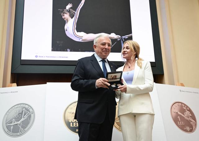 Romanian gymnastics legend Nadia Comaneci (R) and National Bank governor Mugur Isarescu (L) poses next to a prints showing commemorative coins during a ceremony at the National Bank of Romania (BNR) in Bucharest on May 28, 2026. The central bank launched a special numismatic issue—comprising gold, silver, and copper-plated tombac coins—to mark the 50th anniversary of Com?neci achieving the first-ever perfect 10.0 score in Olympic history at the Montreal 1976 Games. The reverse of the coins depicts her iconic uneven bars routine and the historic "1.00" scoreboard display. (Photo by Daniel MIHAILESCU / AFP)