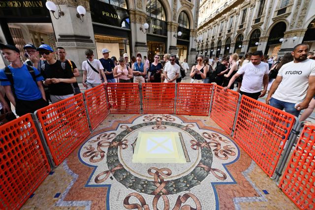 People stop to look at the mosaic of the bull covered for restoration in the Galleria Vittorio Emanuele II, donwtown Milan, on May 27, 2026. "Because of constant pirouettes on the heel made by tourists visiting Milan, the pink tesserae that make up its testicles have been worn down, forming a small crater," city authorities said in a statement. The beige bull mosaic representing Turin, the then capital of Italy, is part of the flooring in the 19th century shopping arcade near Milan's famous Duomo cathedral. (Photo by Stefano RELLANDINI / AFP)