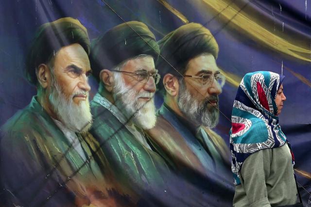 TOPSHOT - An Iranian woman walks past a banner bearing portraits of the late founder of the Islamic Republic, Ayatollah Ruhollah Khomeini (L), former supreme leader Ayatollah Ali Khamenei (C), who was assassinated in February 2026, and current supreme leader Mojtaba Khamenei (R), in Tehran on May 28, 2026. Iran's foreign ministry condemned on May 28, what it called violations by the United States after its strikes on the southern port city of Bandar Abbas and expressed solidarity with Oman after the US president threatened to "blow them up". (Photo by AFP) / 