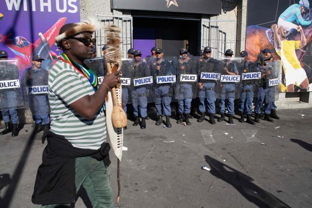 A protester carrying a traditional shield walks past South African Police Service officers during a protest against illegal immigrants organised by March and March in Goodwood, a Cape Town suburb, on May 30, 2026. (Photo by RODGER BOSCH / AFP)