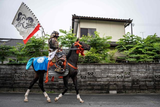 A horseman clad in samurai armor rides through a residential neighbourhood as people gather to watch during the Soma Nomaoi festival in Minamisoma, Fukushima prefecture on May 24, 2026. The Soma Nomaoi began as a way to train mounted warriors and it still looks the same a millennium later, with riders dressed in samurai armour battling it out in horseback events. (Photo by Yuichi YAMAZAKI / AFP) / To go with 'Japan-Climate-Festival-Tradition' Focus by Andrew MCKIRDY
