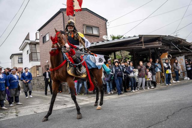 A horseman clad in samurai armor rides through a residential neighbourhood as people gather to watch during the Soma Nomaoi festival in Minamisoma, Fukushima prefecture on May 24, 2026. The Soma Nomaoi began as a way to train mounted warriors and it still looks the same a millennium later, with riders dressed in samurai armour battling it out in horseback events. (Photo by Yuichi YAMAZAKI / AFP) / To go with 'Japan-Climate-Festival-Tradition' Focus by Andrew MCKIRDY