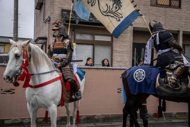 This picture taken on May 24, 2026 shows women looking at horsemen clad in samurai armor as they ride through a residential neighbourhood during the Soma Nomaoi festival in Minamisoma, Fukushima prefecture. The Soma Nomaoi began as a way to train mounted warriors and it still looks the same a millennium later, with riders dressed in samurai armour battling it out in horseback events. (Photo by Yuichi YAMAZAKI / AFP)
