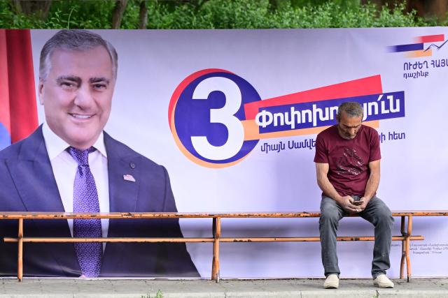 A man sits of a bench next to a campaign poster and a banner of Strong Armenia, the party of billionaire Russian-Armenian businessman Samvel Karapetyan - who is under house arrest on charges of plotting a coup, in Yerevan on June 4, 2026, days ahead of the parliamentary elections. (Photo by Karen MINASYAN / AFP)