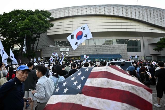 A US flag umbrella is pictured in the crowd as protesters call for the suspension of ballot counting due to a shortage of ballot papers outside a counting station in Seoul on June 5, 2026. South Korean riot police cleared protesters from a Seoul polling station on June 5, after a 35-hour blockade sparked by a ballot shortage during local elections that also led the national election chief to resign. (Photo by Pedro PARDO / AFP)