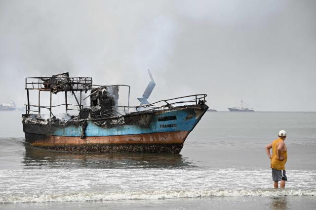 A man stands next to a burned-out fishing boat near the port of Manta, Ecuador on June 6, 2026. At least 35 fishing boats were destroyed and two people were injured in a major fire that broke out at a crowded dock in western Ecuador on June 6, 2026, according to the Risk Management Secretariat. The Ecuadorian Police stated in a press release that "the incident appears to have occurred while welding work was being carried out on a boat" and ruled out the possibility that it was an attack by criminal groups. (Photo by ARIEL OCHOA / AFP)