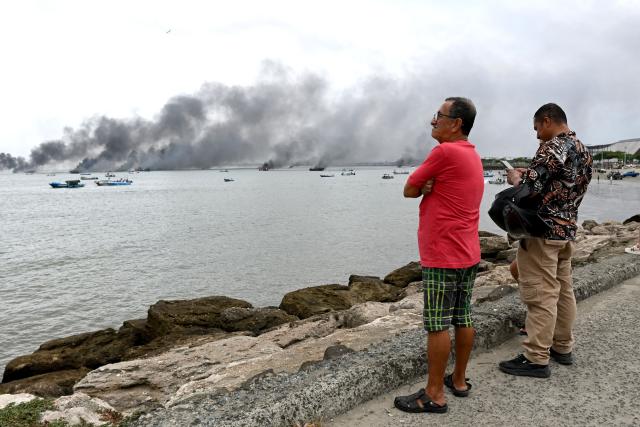 People stand on the coastline as fishing boats burn near the port of Manta, Ecuador on June 6, 2026. At least 35 fishing boats were destroyed and two people were injured in a major fire that broke out at a crowded dock in western Ecuador on June 6, 2026, according to the Risk Management Secretariat. The Ecuadorian Police stated in a press release that "the incident appears to have occurred while welding work was being carried out on a boat" and ruled out the possibility that it was an attack by criminal groups. (Photo by ARIEL OCHOA / AFP)