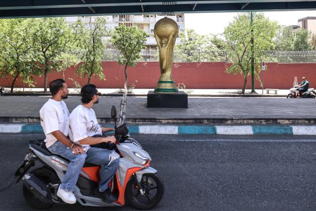 Men ride a scooter past a World Cup trophy displayed under a bridge in Tehran on June 7, 2026. Iran's national team, also known as Team Melli, open their 2026 World Cup campaign, co-hosted by the United States, Mexico and Canada, against New Zealand in Los Angeles on June 15 before lining up against Belgium and Egypt in Group G. Iran has been at war with the United States and Israel since February 28, though a fragile ceasefire has been in place since April 8. Tehran and Washington have had no diplomatic relations since 1980 following the hostage crisis at the US embassy, which came after the Islamic revolution that toppled the US-backed shah. (Photo by ATTA KENARE / AFP)