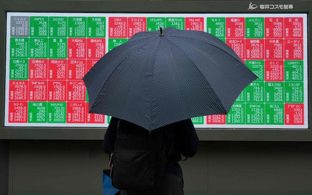 A pedestrian holding an umbrealla stands in front of an electronic quotation board displaying the Nikkei 225 stock prices on the Tokyo Stock Exchange in Tokyo on June 8, 2026. (Photo by Kazuhiro NOGI / AFP)