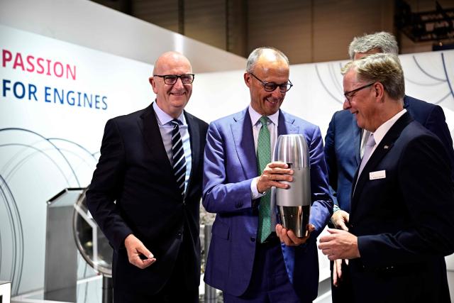 German Chancellor Friedrich Merz (2nd L) chats with Johannes Bussmann, CEO of MTU Aero Engines AG (R) and Brandenburg's State Premier Dietmar Woidke (L) near an aircraft turbine at the booth of MTU Aero Engines AG during his visit at the International Aerospace Exhibition (ILA) in Schoenefeld, near Berlin on June 10, 2026. (Photo by Tobias SCHWARZ / AFP)