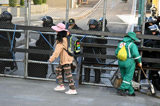 A woman walks past as a sanitation worker sweeps near a riot police barricade during a protest against the government of Bolivia's President Rodrigo Paz in La Paz on June 10, 2026. For weeks, protesters demanding that US-backed conservative President Rodrigo Paz's fledgling government address a deep economic crisis, while others called for his resignation, have marched and set up roadblocks, bringing Bolivia to a near standstill, driving up food and fuel prices and causing shortages of basic goods. (Photo by AIZAR RALDES / AFP)