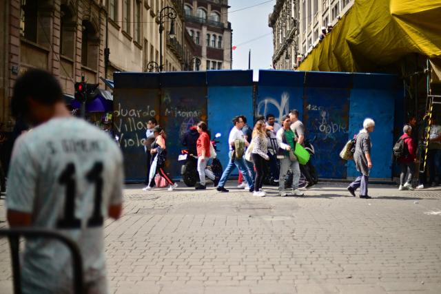 People walk past a security barrier set up to block access to the Fan Fest at Mexico City's Zocalo square on June 10, 2026, amid a threatened protest by the National Coordinator of Education Workers (CNTE) ahead of the 2026 FIFA World Cup. (Photo by Marco Antonio MARTINEZ / AFP)