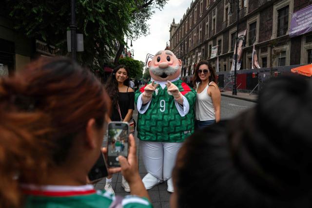 Tourists take a photo with a Dr. Simi character wearing a Mexican football jersey near the FIFA Fan Fest at Zocalo Square, one day before the opening of the 2026 FIFA World Cup football tournament in Mexico City on June 10, 2026. (Photo by Gerardo Luna / AFP)