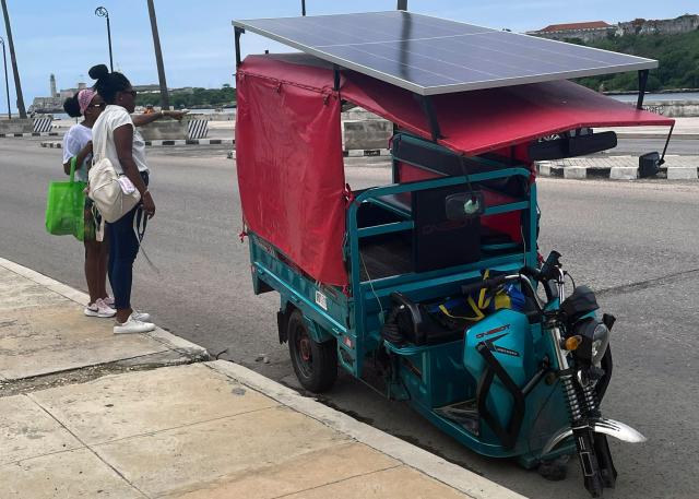 Women stand next to a broken-down tricycle fitted with a solar panel to recharge its battery in the Old Havana neighborhood in Havana on June 11, 2026. (Photo by Pablo PORCIUNCULA / AFP)