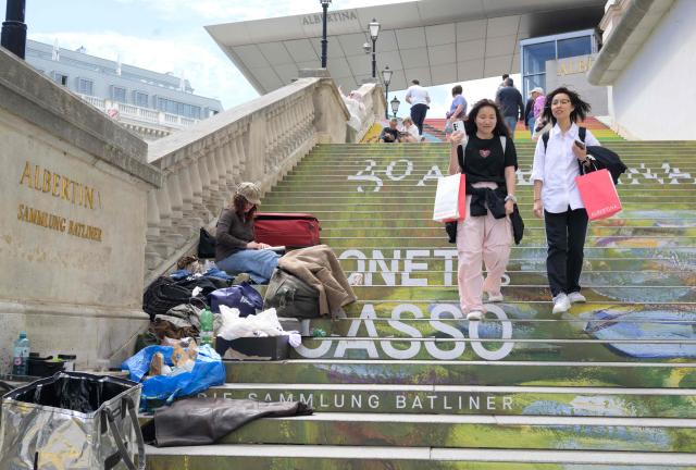 A homeless woman camps on the left side of a stairway to the famous Albertina art gallery while visitors pass through the other side, in Vienna, Austria, on June 12, 2026. (Photo by Joe Klamar / AFP)