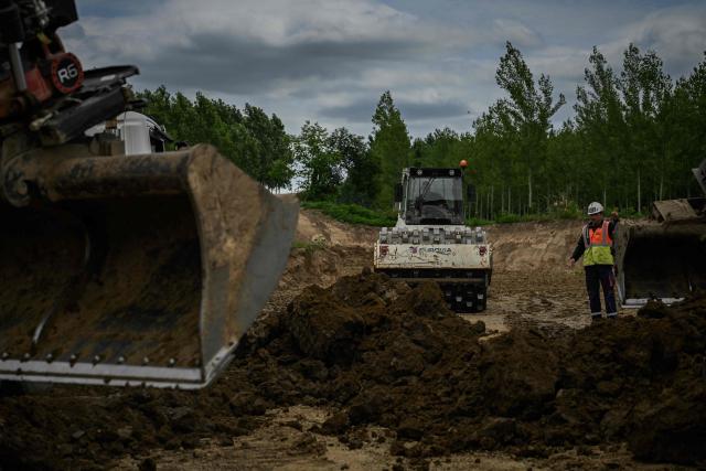 A construction worker gestures as a sheepfoot compactor roller packs clay on a dyke reconstruction site along the Garonne river in Senestis, south-western France, on June 8, 2026. In Lot-et-Garonne, more than a kilometer of dikes broken by the February floods -its biggest flood in two decades- are being rebuilt, at a cost estimated at eight million euros which is weighing on local finances. To fill the hole, 25 trucks transport approximately 2,000 tons of a special clay every day. The work is expected to continue until the fall. (Photo by Philippe LOPEZ / AFP)