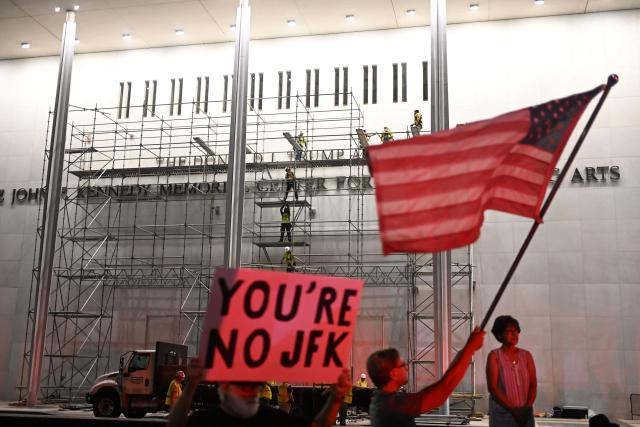 Protesters wave a US flag and hold a sign reading "you're no JFK" as workers build scaffolding near the signage for the "Donald J. Trump and John F. Kennedy Memorial Center for the Performing Arts" in Washington, DC, on June 12, 2026. A US federal judge on June 12 rejected a bid by the board of the Kennedy Center and the Justice Department to halt the removal of President Donald Trump's name from the performing arts venue. District Judge Christopher Cooper, in a ruling last month, ordered Trump's name taken off the iconic building in the nation's capital by Friday, June 12, 2026. (Photo by Alex WROBLEWSKI / AFP)