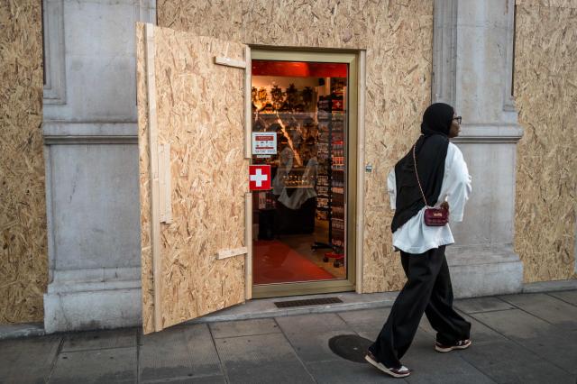 A tourist walks past a souvenir shop, with its door adorned with a Swiss cross, boarded up with wooden panels to protect the façade ahead of demonstrations against the nearby G7 summit in Evian, in Geneva, June 13, 2026. Protests against the G7 summit in Evian are set to take place in nearby Geneva, where activists can gather more easily than in the tightly secured host town. (Photo by Fabrice COFFRINI / AFP)