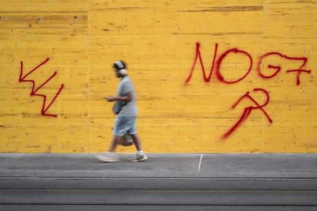 A passer-by walks past a shop boarded up with graffiti-covered wooden panels to protect the façade ahead of demonstrations against the nearby G7 summit in Evian, in Geneva, June 13, 2026. Protests against the G7 summit in Evian are set to take place in nearby Geneva, where activists can gather more easily than in the tightly secured host town. (Photo by Fabrice COFFRINI / AFP)