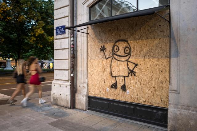 Passers-by walks past a building boarded up with graffiti-covered wooden panels to protect the façade ahead of demonstrations against the nearby G7 summit in Evian, in Geneva, June 12, 2026. Protests against the G7 summit in Evian are set to take place in nearby Geneva, where activists can gather more easily than in the tightly secured host town. (Photo by Fabrice COFFRINI / AFP)
