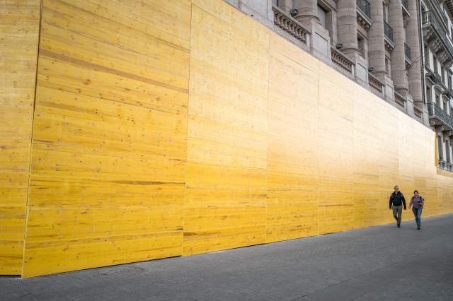 Passers-by walk past along a building boarded up with wooden panels to protect the façade ahead of demonstrations against the nearby G7 summit in Evian, in Geneva, June 13, 2026. Protests against the G7 summit in Evian are set to take place in nearby Geneva, where activists can gather more easily than in the tightly secured host town. (Photo by Fabrice COFFRINI / AFP)