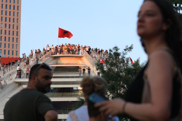 Protestors atop a building hold national flags as they gather in front of Albania's Prime Minister's Office, in Tirana, on June 13, 2026 during a demonstration against the construction of a luxury resort near a protected natural area of Zvernec near Vlore. Several thousand citizens demonstrated in Tirana, 14 days in row after the first protest  repressed on Zvernec beach, to demand transparency around a tourist complex project allegedly linked to US President's son-in-law, Jared Kushner. The demonstrators argue that the estimated $1.2 billion project, led by Kushner and his wife Ivanka, will harm the environment, with plans for luxury hotels in the protected area of Vjosa-Narta on the southern coast. (Photo by Adnan Beci / AFP)
