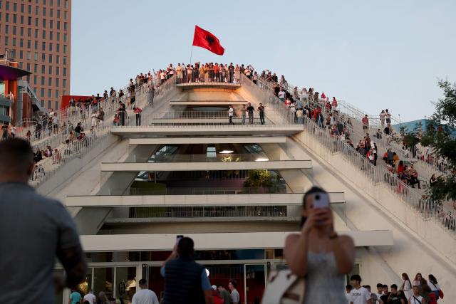 Protestors atop a building hold national flags as they gather in front of Albania's Prime Minister's Office, in Tirana, on June 13, 2026 during a demonstration against the construction of a luxury resort near a protected natural area of Zvernec near Vlore. Several thousand citizens demonstrated in Tirana, 14 days in row after the first protest  repressed on Zvernec beach, to demand transparency around a tourist complex project allegedly linked to US President's son-in-law, Jared Kushner. The demonstrators argue that the estimated $1.2 billion project, led by Kushner and his wife Ivanka, will harm the environment, with plans for luxury hotels in the protected area of Vjosa-Narta on the southern coast. (Photo by Adnan Beci / AFP)