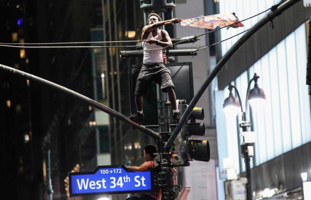 A Knicks fan waves a flag while standing on top of a traffic light poles after the New York Knicks defeated the San Antonio Spurs in Game 5 of the NBA Finals to win the series, in New York, on June 14, 2026. The Knicks won the NBA Championship for the first time since 1973, for the third time in franchise history. (Photo by kena betancur / AFP)