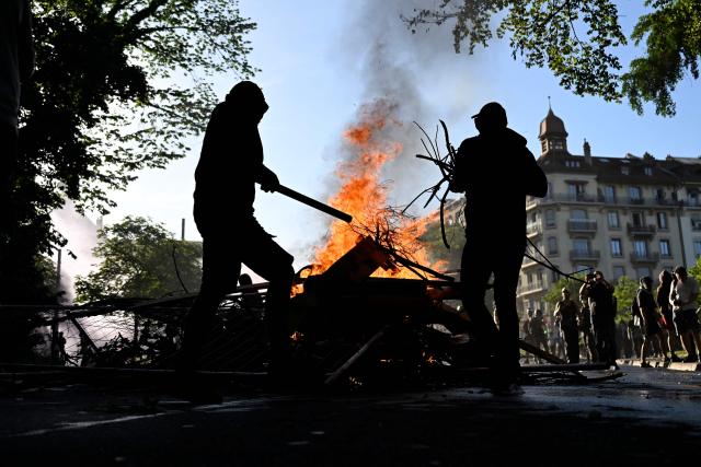 Demonstrators place wood and branches in the fire of a burning trash container during a rally of a "No-G7" coalition of over 60 associations, unions and left-wing groups "against fascism and imperialism, a day ahead of the summit's start, in Geneva on June 14, 2026. The 2026 edition of the G7 summit is set to take place June 15 to 17 in the French town of Evian-les-Bains near Switzerland, attended by country leaders as well as the EU's foreign policy chief and ministers from Brazil, Canada, the United Arab Emirates and Turkey. (Photo by Gabriel Monnet / AFP)