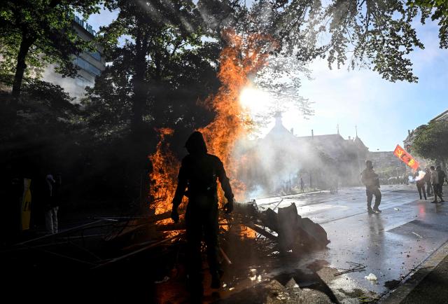 A demonstrator stands next to the fire of a burning trash container during a rally of a "No-G7" coalition of over 60 associations, unions and left-wing groups "against fascism and imperialism, a day ahead of the summit's start, in Geneva on June 14, 2026. The 2026 edition of the G7 summit is set to take place June 15 to 17 in the French town of Evian-les-Bains near Switzerland, attended by country leaders as well as the EU's foreign policy chief and ministers from Brazil, Canada, the United Arab Emirates and Turkey. (Photo by Gabriel Monnet / AFP)