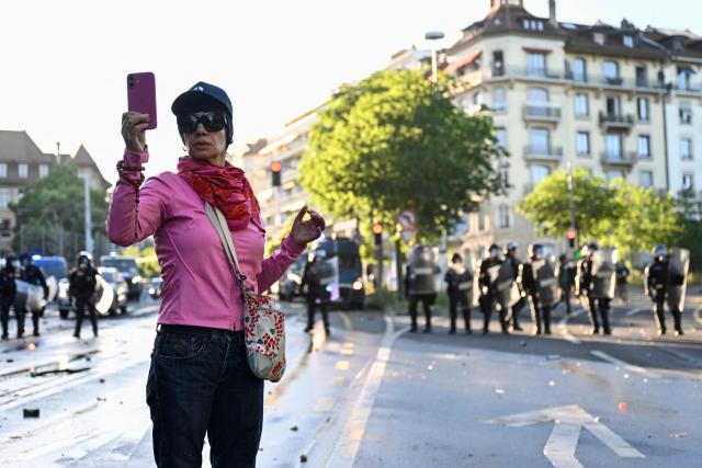 A woman with sunglasses holds a smartphone as riot police operate in the background during a rally of a "No-G7" coalition of over 60 associations, unions and left-wing groups "against fascism and imperialism, a day ahead of the summit's start, in Geneva on June 14, 2026. The 2026 edition of the G7 summit is set to take place June 15 to 17 in the French town of Evian-les-Bains near Switzerland, attended by country leaders as well as the EU's foreign policy chief and ministers from Brazil, Canada, the United Arab Emirates and Turkey. (Photo by Gabriel Monnet / AFP)