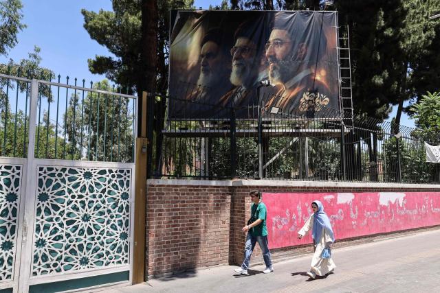 Iranians walk past a billboard depicting Iran's late supreme leader Ayatollah Ruhollah Khomeini (L), his successor, the late Ayatollah Ali Khamenei (C), and the new supreme leader Mojtaba Khamenei outside the former US embassy in Tehran on June 15, 2026. The United States and Iran agreed a peace deal and an "immediate and permanent" end to military operations on all fronts including Lebanon, mediator Pakistan said, signaling the apparent end to more than three months of war in Middle East. (Photo by ATTA KENARE / AFP) / 
