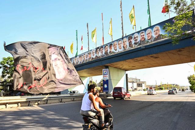 A man holds a huge flag with the image of former Shia Muslim Hezbollah chief Hassan Nasrallah, who was killed in massive Israeli strikes on Beirut's southern suburbs in September 2024 during a previous round of hostilities, as he ride pillion along the highway past the coastal town of Ghazieyh, as displaced families return to their home villages in southern Lebanon on June 15, 2026. Israel's defence minister said on June 15 that Israeli forces would remain in Lebanon, Syria and Gaza indefinitely, hours after the United States and Iran agreed to end the Middle East war, including in Lebanon.  Lebanon was drawn into the Middle East war on March 2 when Tehran-backed militant group Hezbollah launched attacks on Israel to avenge the killing of the Iranian leader. Israel has responded with broad strikes across Lebanon and a ground offensive. (Photo by FADEL itani / AFP) / 