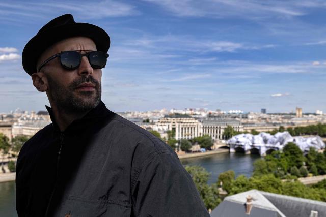 French artist JR looks on with his latest artwork in the background, the Pont-Neuf bridge displaying "La Caverne du Pont Neuf" ("The Pont Neuf Cave") in Paris on June 1, 2026. (Photo by JOEL SAGET / AFP)