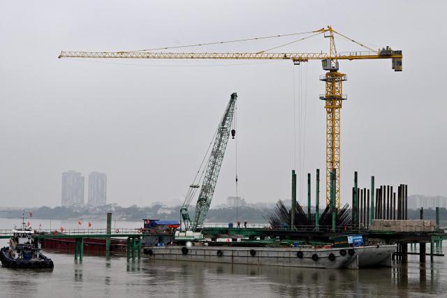 Workers use heavy construction equipment to build a new bridge over the Red River in Hanoi on June 16, 2026. (Photo by Nhac NGUYEN / AFP)