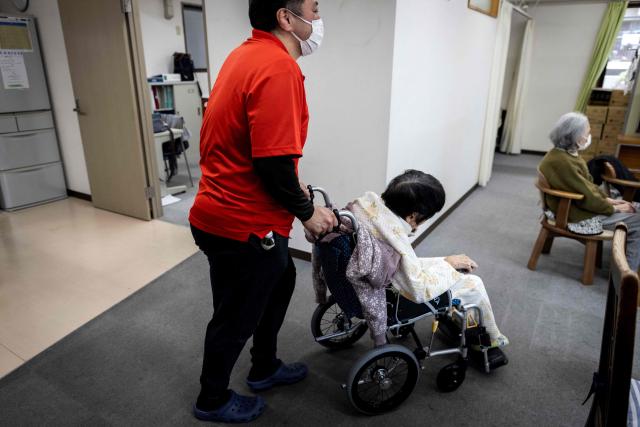 (FILES) A caretaker wheels an elderly woman after a bath at a day care facility for senior citizens in Tokyo on April 6, 2022. In Yo Kusakabe's taboo novel, a young doctor advocates chopping off elderly patients' useless limbs as a groundbreaking solution to super-ageing Japan's overstressed care industry. Now adapted into a film, "Haiyoshin (Useless Body)" has attracted huge controversy since its release last month in Japan, but has also focused attention on the struggling care sector in a country with the world's second-oldest population. (Photo by Behrouz MEHRI / AFP)