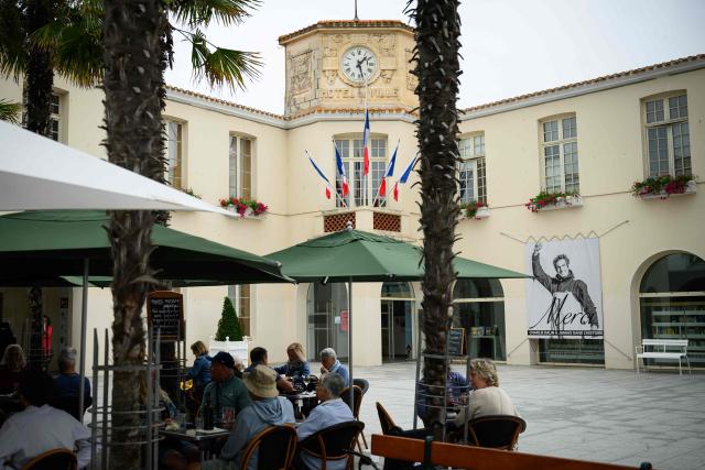 People eat in a terrasse in front of flags at half-staff and a poster in memory of late French skipper Charlie Dalin displayed in front of the city hall in Les Sables-d'Olonne, western France on June 16, 2026. French offshore sailor and Vendee Globe 2024/2025 winner Charlie Dalin has died in Quimper, western France, aged 42, from cancer, his family announced on June 11, 2026. (Photo by Loic VENANCE / AFP)