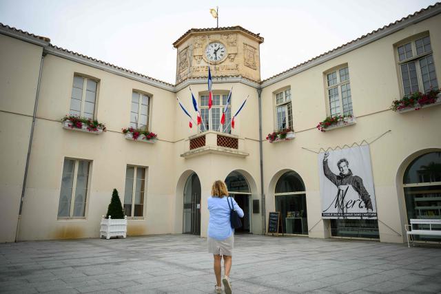 A woman walks in front of flags at half-staff and a poster in memory of late French skipper Charlie Dalin displayed on the facade of the city hall in Les Sables-d'Olonne, western France on June 16, 2026. French offshore sailor and Vendee Globe 2024/2025 winner Charlie Dalin has died in Quimper, western France, aged 42, from cancer, his family announced on June 11, 2026. (Photo by Loic VENANCE / AFP)