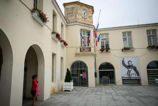 A woman stands next to flags at half-staff and a poster in memory of late French skipper Charlie Dalin displayed in front of the city hall in Les Sables-d'Olonne, western France on June 16, 2026. French offshore sailor and Vendee Globe 2024/2025 winner Charlie Dalin has died in Quimper, western France, aged 42, from cancer, his family announced on June 11, 2026. (Photo by Loic VENANCE / AFP)