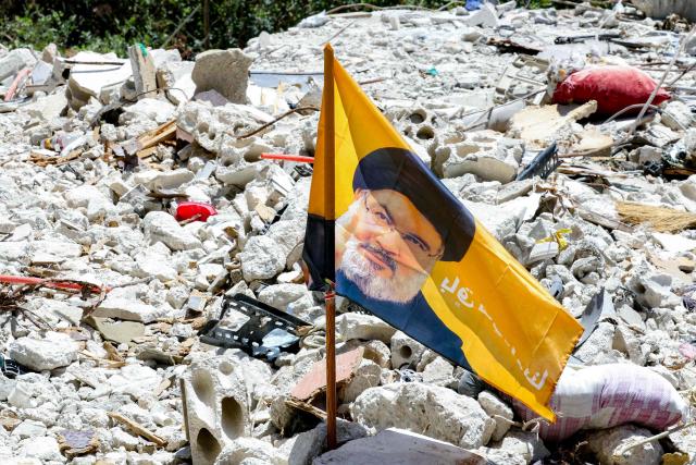 A flag bearing the image of Hezbollah slain leader Hassan Nasrallah, who was killed by an Israeli airstrike on Beirut's southern suburbs in 2024, is placed amidst rubble in the village of Deir Qanoun al-Nahr in southern Lebanon on June 16, 2026. Lebanese state media said that Israeli strikes killed at least four people in the Nabatieh area of the country's south on June 16, despite an announcement that a US-Iran deal included the Israel-Hezbollah war. While violence has declined in Lebanon since the agreement between the US and Iran was announced, Israeli strikes on the south have now killed at least five people since June 15. (Photo by MAHMOUD ZAYYAT / AFP) / 