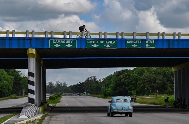 A person rides a bicycle along a bridge with signs pointing to Camaguey, Ciego de Avila and Sancti Spiritus, as an old car rides along the National Motorway, on the outskirts of Havana, on June 16, 2026. New restrictions on interprovincial transport in Cuba come into force on June 18, 2026, with trains running every 16 days, few weekly coaches and a state system for allocating tickets according to criteria of "urgency", amid a severe fuel shortage, under pressure from Washington. (Photo by YAMIL LAGE / AFP)