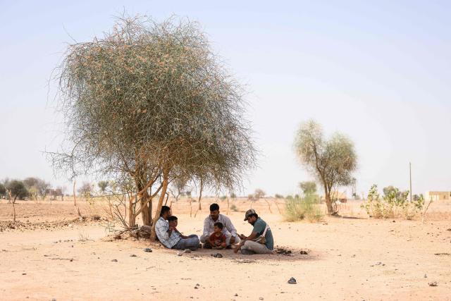 This photograph taken on May 29, 2026 shows herdsman Sawai Singh Bhati (C) sitting with his son under the shade of a tree in 44 degrees Celsius temperatures, near his house in Sanwata village on the outskirts of Rajasthan's Jaisalmer city. In India's hot plains, scorching summers have become increasingly harder to endure, requiring adaptations and forcing life into the hours of dark before the sun turns punishing. Temperatures in Singh's Sanwata village hit 45C in early June, as is often during the summer with the highest temperature recorded in the area as 49C. (Photo by Manan VATSYAYANA / AFP)