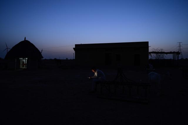 This photograph taken on May 29, 2026 shows herdsman Sawai Singh Bhati checking his mobile phone in 34 degrees Celsius temperatures, outside his house after returning from work, in Sanwata village on the outskirts of Rajasthan's Jaisalmer city. In India's hot plains, scorching summers have become increasingly harder to endure, requiring adaptations and forcing life into the hours of dark before the sun turns punishing. Temperatures in Singh's Sanwata village hit 45C in early June, as is often during the summer with the highest temperature recorded in the area as 49C. (Photo by Manan VATSYAYANA / AFP)