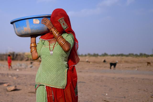 This photograph taken on May 29, 2026 shows shows herdsman Sawai Singh Bhati's mother, Seema Kawar carrying a plastic pan after feeding cattle outside her house in Sanwata village on the outskirts of Rajasthan's Jaisalmer city. In India's hot plains, scorching summers have become increasingly harder to endure, requiring adaptations and forcing life into the hours of dark before the sun turns punishing. Temperatures in Singh's Sanwata village hit 45C in early June, as is often during the summer with the highest temperature recorded in the area as 49C. (Photo by Manan VATSYAYANA / AFP)