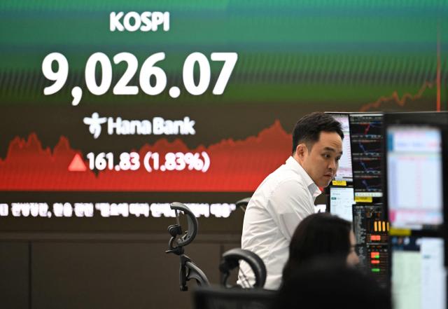 Currency dealers monitor exchange rates as a screen shows South Korea's benchmark stock index (KOSPI) as it breaks 9,000 points in a foreign exchange dealing room at the Hana Bank headquarters in Seoul on June 18, 2026. South Korea's benchmark Kospi index topped 9,000 points for the first time on June 18, on the back of a tech-fuelled rally led by chip titans Samsung and SK hynix. (Photo by Jung Yeon-je / AFP)