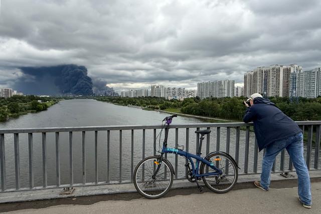 A man takes photographs from a bridge across the Moskva river as black smoke rises from the area of the Russian oil producer Gazprom Neft's Moscow oil refinery on the south-eastern outskirts of Moscow on June 18, 2026. Moscow was fending off a "large-scale" drone attack from Ukraine, with several drones reaching an oil refinery, the city's mayor Sergey Sobyanin said early on June 18, 2026. (Photo by AFP)