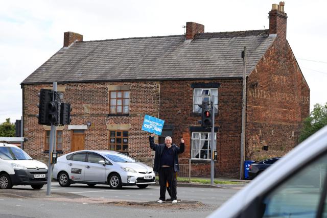 A person holds a placard in support of the Reform UK party in the middle of a junction in Winstanley, part of the Makerfield parliamentary constituency, in north west England on June 18, 2026, as voters go to the polls to vote in the Makerfield by-election. Britain holds a unique vote on June 18 that could trigger the endgame for Labour leader Keir Starmer's beleaguered premiership, or win him a reprieve. (Photo by Darren Staples / AFP)