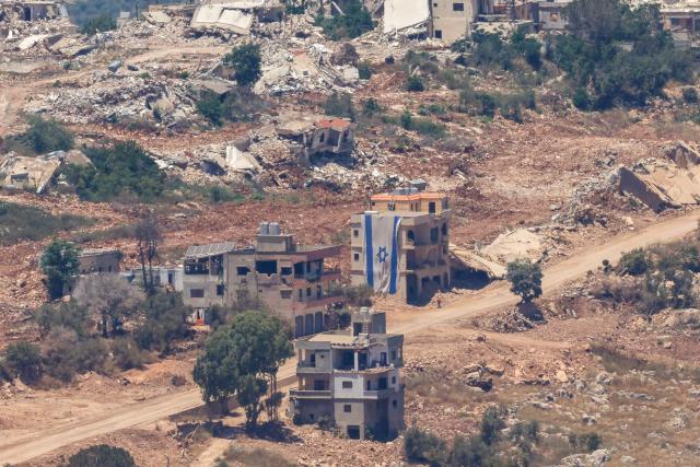 This picture taken from a position in the Upper Galilee in northern Israel, a man walking past an Israeli flag covering part of a destroyed building in the southern Lebanese village of Taybeh near the border on June 18, 2026. Israeli strikes in south Lebanon killed three people on June 18, according to Lebanese state media, hours after the United States and Iran signed an agreement to end the Middle East war. Israel's military, meanwhile, announced the death of one of its soldiers the night before in an incident in south Lebanon that also left seven others wounded. (Photo by Jalaa MAREY / AFP)