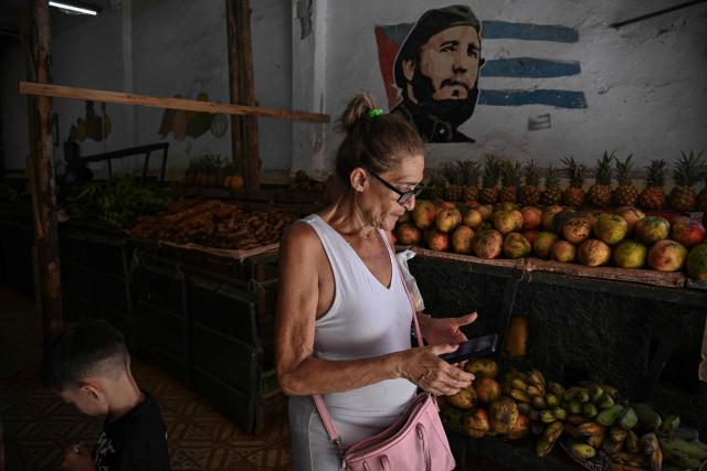 A woman checks her mobile phone at a farmers’ market in Havana on June 18, 2026, past a photograph of late Cuban leader Fidel Castro. (Photo by YAMIL LAGE / AFP)