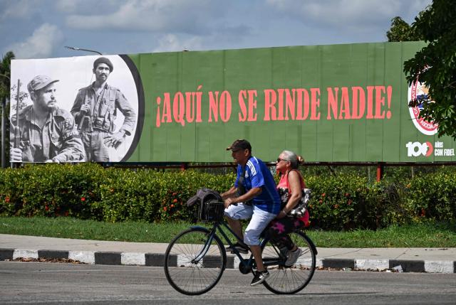 A man and a woman ride a bicycle past a sign featuring the image of Cuba's late leader Fidel Castro (1926-2016) and commander Juan Almeida Bosque (1927-2009) reading “Nobody surrenders here”, in Havana on June 19, 2026. Nearly 70 years after Fidel Castro's 1959 revolution, communist-run Cuba has adopted a sweeping package of free-market reforms aimed at extricating the country from a deep crisis. (Photo by YAMIL LAGE / AFP)